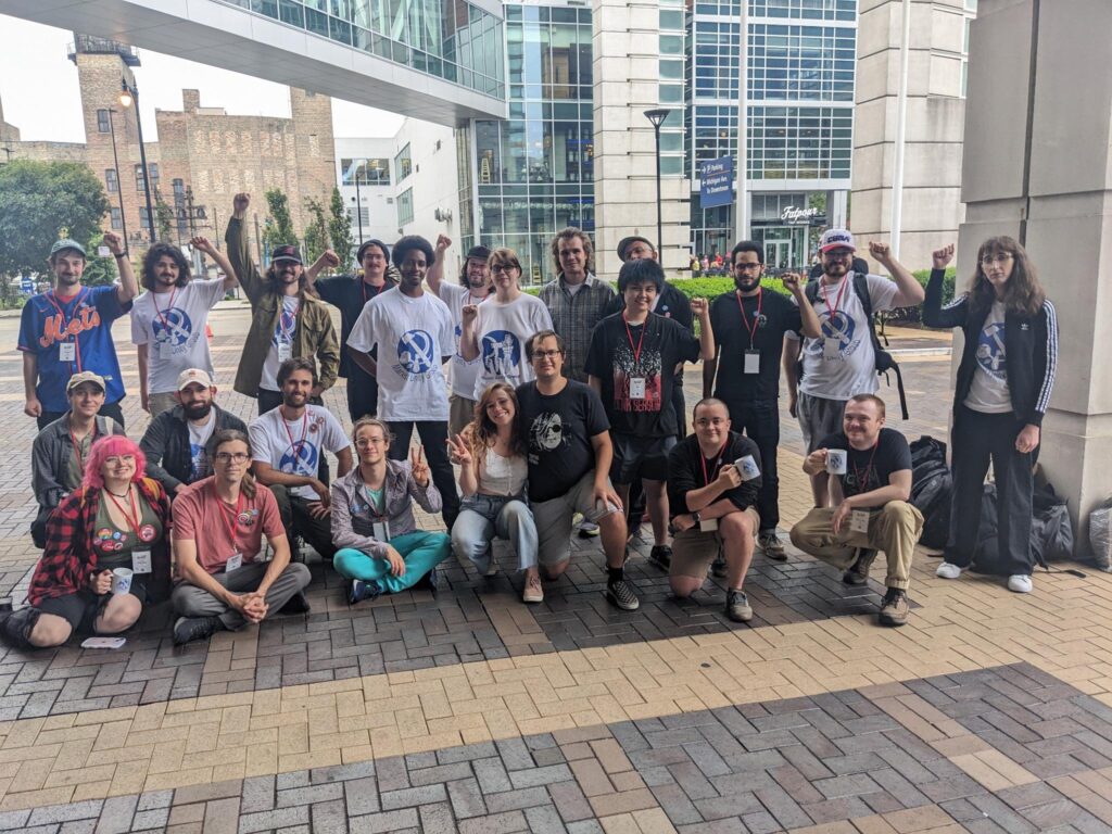 Marxist Unity Group members pose for a group photo in front of the hotel where the 2023 DSA convention took place. Folks are wearing "Marxist Unity Group" shirts and holding Marxist Unity Group MUGs.