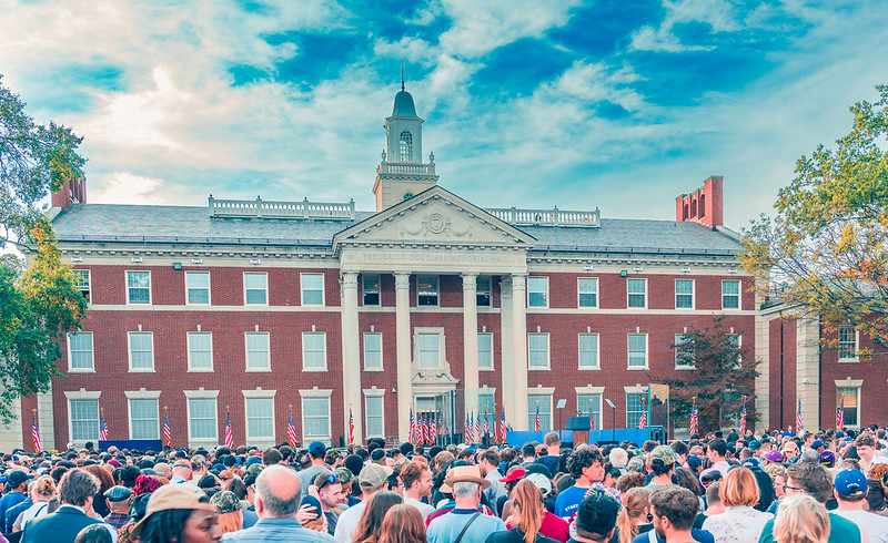 Crowd of people standing in front of Howard University's Douglass hall waiting for Kamala Harris to give her concession speech. An empty podium surrounded by American flags is in front of the crowd waiting.